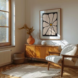 May include: A room interior featuring a framed floral art print with a black background and cream-colored petals. The room includes a wooden cabinet, a woven basket, a beige armchair, and a terracotta vase with dried foliage. A rug and a window are also visible.