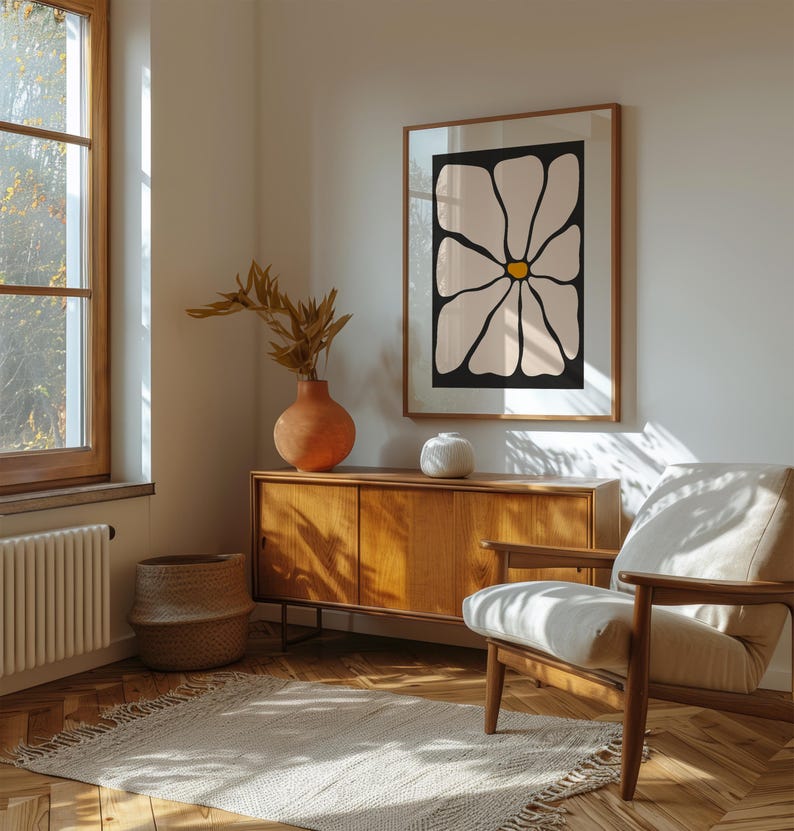 May include: A room interior featuring a framed floral art print with a black background and cream-colored petals. The room includes a wooden cabinet, a woven basket, a beige armchair, and a terracotta vase with dried foliage. A rug and a window are also visible.