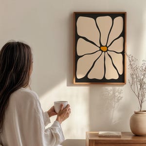May include: A framed wall art print featuring a large, stylized daisy flower with cream-colored petals and a yellow center against a black background. The artwork is in a dark brown wooden frame. The image is a minimalist, modern design.