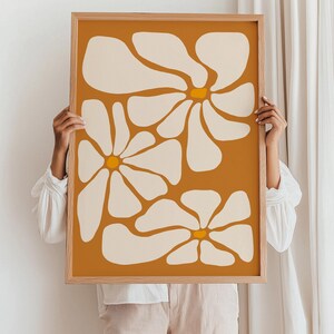 May include: A framed print with a minimalist design featuring three stylized white flowers with yellow centers on a mustard yellow background.