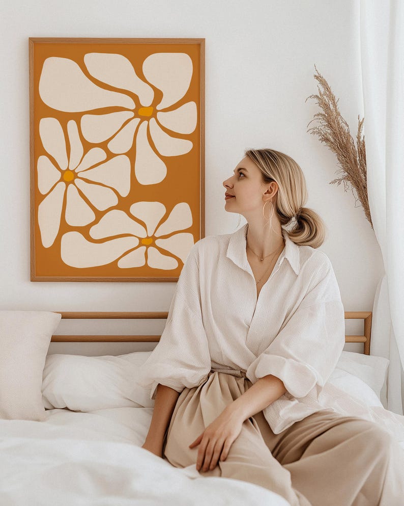 May include: A framed print with a minimalist design of white flowers on a burnt orange background. The print is hanging on a white wall above a bed with white bedding. A person is sitting on the bed wearing a white shirt and beige pants.