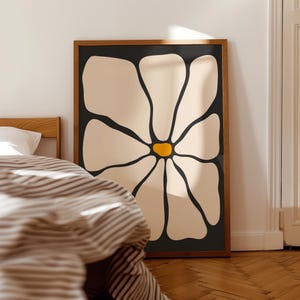 May include: A framed art print featuring a large, stylized flower. The flower has cream-colored petals with black outlines and a yellow center, set against a black background. The wooden frame adds a natural touch. The artwork is leaning against a wall.