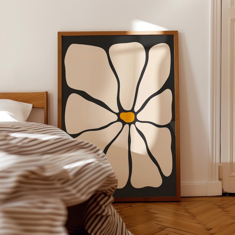 May include: A framed art print featuring a large, stylized flower. The flower has cream-colored petals with black outlines and a yellow center, set against a black background. The wooden frame adds a natural touch. The artwork is leaning against a wall.
