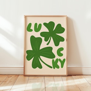 Puede incluir: Un estampado artístico enmarcado con la palabra "LUCKY" en letras verdes y tres tréboles verdes. El fondo es de color beige claro. El marco es de color marrón claro.