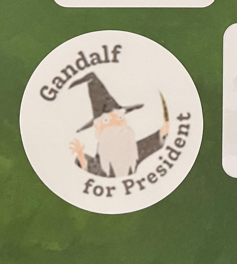 May include: A white circle sticker with a cartoon image of Gandalf from the Lord of the Rings, wearing a black pointed hat and holding a staff. The text "Gandalf for President" is written around the image.