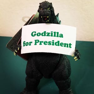 May include: A dark green Godzilla action figure with textured skin and yellow claws, holding a white rectangular sign that features the green text "Godzilla for President" in a bold, sans-serif font. The figure has an open mouth.