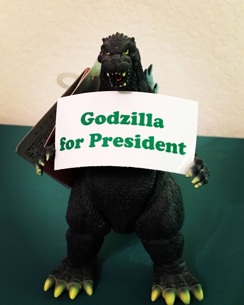 May include: A dark green Godzilla action figure with textured skin and yellow claws, holding a white rectangular sign that features the green text "Godzilla for President" in a bold, sans-serif font. The figure has an open mouth.
