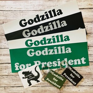 May include: A collection of rectangular stickers and keychains featuring the text Godzilla and Godzilla for President. The stickers are white, black, and green with contrasting text. Two rectangular keychains in green and black display the text Godzilla for President, and one square white sticker shows a black silhouette of Godzilla with the text Godzilla for President.