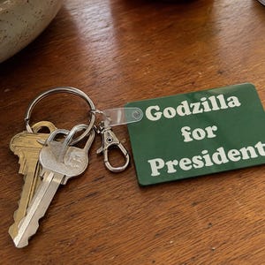 May include: A rectangular green keychain tag with white text that reads Godzilla for President. The tag is attached to a silver metal split ring holding two keys, one brass and one silver, and a small silver lobster claw clasp connector.