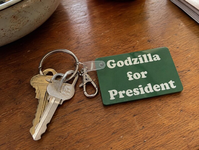 May include: A rectangular green keychain tag with white text that reads Godzilla for President. The tag is attached to a silver metal split ring holding two keys, one brass and one silver, and a small silver lobster claw clasp connector.