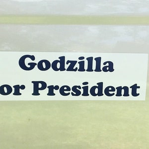 May include: A rectangular white sticker featuring the text "Godzilla for President" printed in a bold, dark blue, serif font. The sticker is presented against a light, neutral background, highlighting the clear and legible lettering.