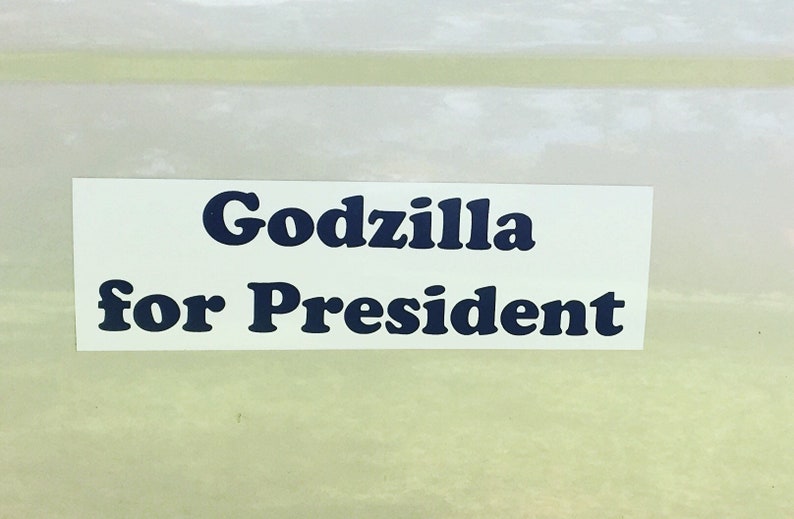 May include: A rectangular white sticker featuring the text "Godzilla for President" printed in a bold, dark blue, serif font. The sticker is presented against a light, neutral background, highlighting the clear and legible lettering.
