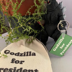 May include: A black Godzilla figurine with grey dorsal fins, a green square keychain with the text Godzilla for President, and a beige canvas drawstring pouch featuring the black printed text Godzilla for President.