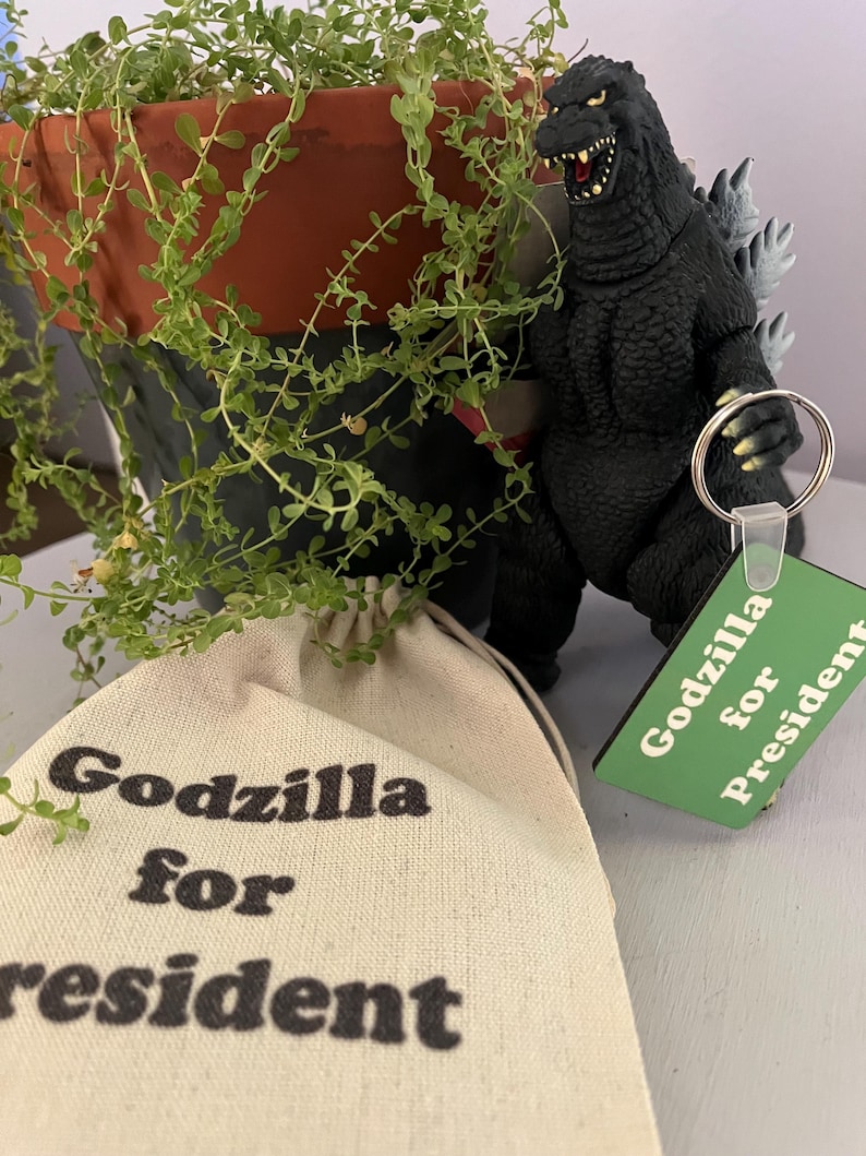 May include: A black Godzilla figurine with grey dorsal fins, a green square keychain with the text Godzilla for President, and a beige canvas drawstring pouch featuring the black printed text Godzilla for President.
