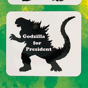 May include: A square white sticker featuring a black silhouette of the monster Godzilla. The text "Godzilla for President" is printed in white, bold, sans-serif capital and lowercase letters across the center of the creature's body.