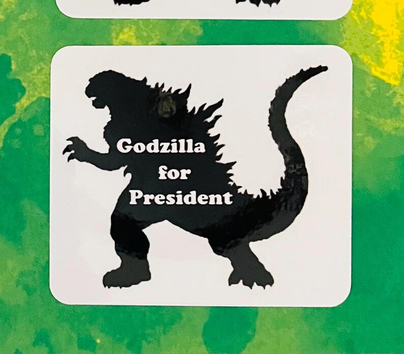 May include: A square white sticker featuring a black silhouette of the monster Godzilla. The text "Godzilla for President" is printed in white, bold, sans-serif capital and lowercase letters across the center of the creature's body.