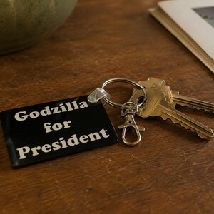 May include: A rectangular black keychain with white text that reads Godzilla for President. The keychain is attached to a silver metal split ring and a lobster claw clasp, with two silver keys connected to the ring.