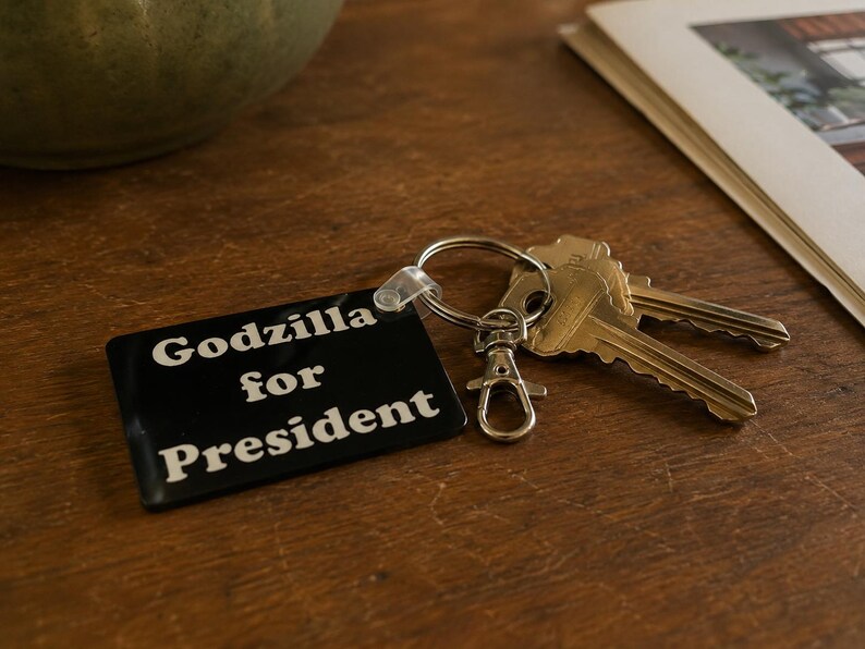 May include: A rectangular black keychain with white text that reads Godzilla for President. The keychain is attached to a silver metal split ring and a lobster claw clasp, with two silver keys connected to the ring.