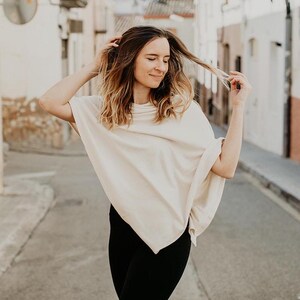 Peut inclure: Une femme portant un poncho cr&egrave;me &agrave; la coupe ample et fluide. Elle se tient dans une rue &eacute;troite avec des b&acirc;timents blancs de chaque c&ocirc;t&eacute;.