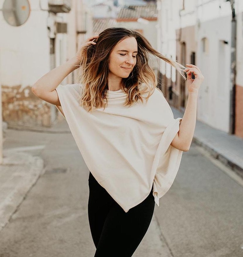 Peut inclure: Une femme portant un poncho cr&egrave;me &agrave; la coupe ample et fluide. Elle se tient dans une rue &eacute;troite avec des b&acirc;timents blancs de chaque c&ocirc;t&eacute;.