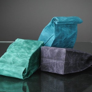 Puede incluir: Tres bolsas de almuerzo reutilizables hechas de lona encerada. Las bolsas est&aacute;n dobladas y son de color turquesa, verde y gris.