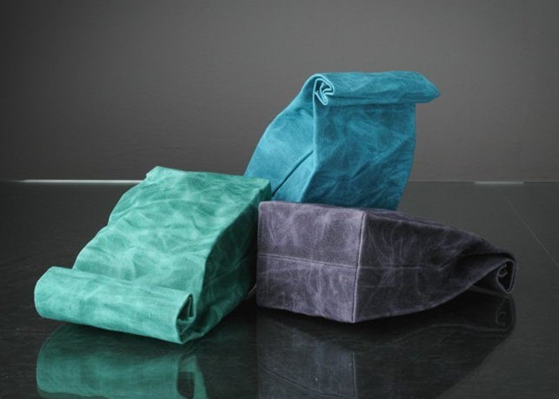 Puede incluir: Tres bolsas de almuerzo reutilizables hechas de lona encerada. Las bolsas est&aacute;n dobladas y son de color turquesa, verde y gris.