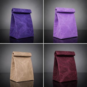 May include: A set of four rectangular lunch bags with a roll-top closure. Each bag features a distinct solid color: deep purple, light lavender, beige, and dark burgundy. The material has a textured, crinkled appearance throughout the surface.
