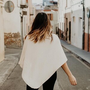 Peut inclure: Une femme portant un poncho blanc et un pantalon noir marche dans une rue &eacute;troite.