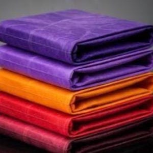 May include: A stack of five folded rectangular fabric pieces in vibrant colors. The top two are purple, followed by an orange piece, a bright red piece, and a dark maroon piece at the bottom. Each piece features a subtle, repeating grid pattern.