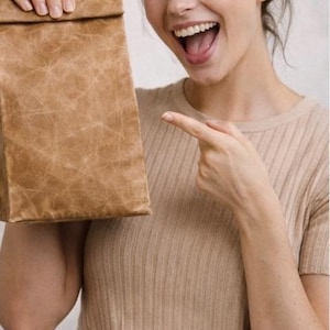 May include: A rectangular, light brown lunch bag with a textured, crinkled surface finish. The bag features a folded top closure and a structured, upright design. The material has a distinct, weathered appearance with a grid-like pattern of creases.