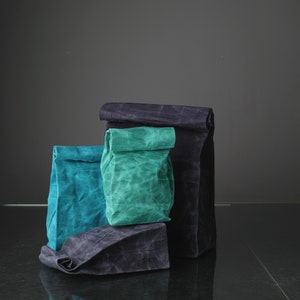 Puede incluir: Tres bolsas de tela reutilizables, una negra, una azul verdosa y una verde azulada, est&aacute;n apiladas una encima de la otra. Las bolsas est&aacute;n hechas de una tela texturizada y tienen la parte superior doblada.