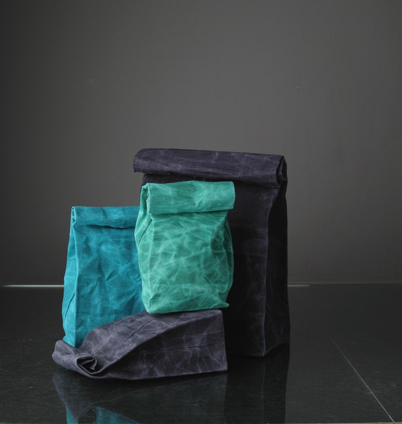 Puede incluir: Tres bolsas de tela reutilizables, una negra, una azul verdosa y una verde azulada, est&aacute;n apiladas una encima de la otra. Las bolsas est&aacute;n hechas de una tela texturizada y tienen la parte superior doblada.