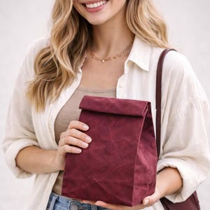 May include: A deep burgundy, rectangular lunch bag with a folded top closure and a textured, paper-like finish. The bag is held in front of a light-colored shirt and features a dark brown shoulder strap attached to the side of the bag.