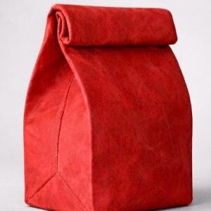 May include: A bright red, rectangular lunch bag with a textured, paper-like surface. The bag features a roll-top closure at the top, creating a folded flap design. The structure is upright and vertical, with visible seams along the front and sides.