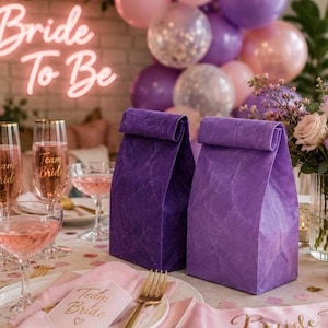 May include: Two upright purple paper bags with a textured finish sit on a table alongside pink champagne glasses, a plate with a Team Bride card, and a neon Bride To Be sign in the background with pink and purple balloons.