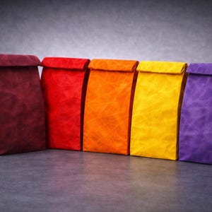 May include: A row of five upright paper bags with folded tops, arranged in a gradient of colors from left to right: deep burgundy, bright red, vibrant orange, sunny yellow, and a rich purple. Each bag features a textured, matte finish.