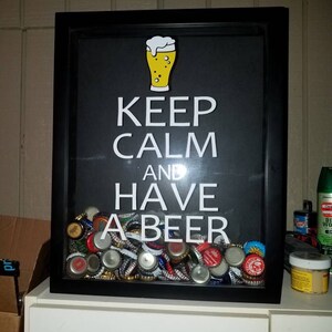 May include: A black framed shadow box with the text "Keep Calm and Have A Beer" in white letters. A yellow beer glass icon is above the text. The box is filled with bottle caps.