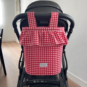 May include: A red and white gingham fabric storage bag attached to the back of a black stroller. The bag features a ruffled top edge, two fabric straps hanging from the stroller handle, and a white rectangular label with the text FOR EMMY.