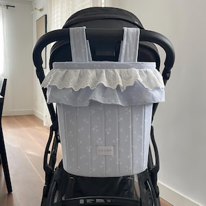 May include: A light blue fabric stroller organizer featuring a white floral pattern, a white eyelet lace ruffle trim along the top edge, and two vertical hanging straps. A rectangular white label with the black text FOR EMMY is centered.