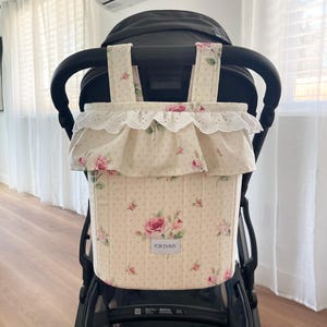 May include: A cream-colored quilted stroller organizer featuring a pink rose floral pattern and small brown polka dots. The bag has two top straps, a ruffled trim with white eyelet lace, and a rectangular label that reads FOR EMMY.