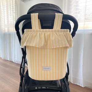 May include: A yellow and white striped stroller organizer bag featuring a ruffled top edge, two vertical hanging straps, and a rectangular fabric label centered on the front with the text FOR EMMY printed in black capital letters.