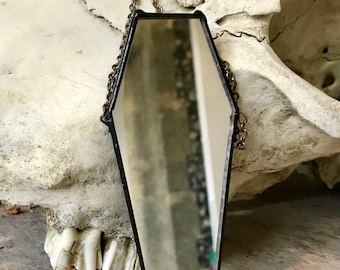 May include: A black coffin-shaped mirror with a silver chain hanging from the top. The mirror is reflecting a blurry image of a skull.