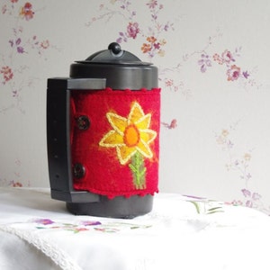 Könnte beinhalten: Eine schwarze isolierte Kaffeekanne mit einer roten Filz-Abdeckung, die eine gestickte gelbe Blume zeigt. Die Blume hat eine gelbe Mitte und einen grünen Stiel.