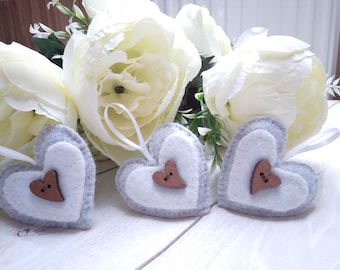May include: Three gray and white felt heart ornaments with wooden buttons. The hearts are hanging from white ribbon.