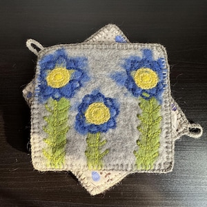May include: A grey wool pot holder with a crocheted border features three embroidered blue flowers with yellow centres and green stems.