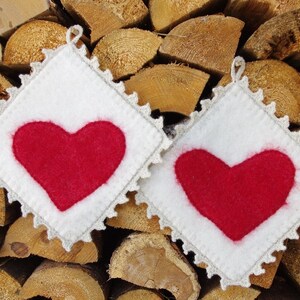 Może przedstawiać: Dwa białe, szydełkowe uchwyty na garnki z czerwonymi sercami z filcu w środku. Uchwyty leżą na tle ułożonych drewnianych kłód.