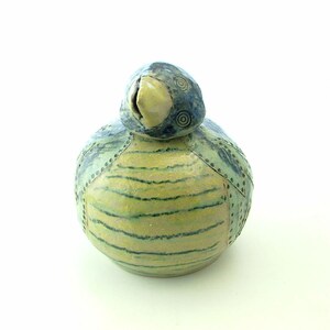 Op de afbeelding: Een keramiek vogelbeeldje met een blauw en groen gestreept lichaam en een gele snavel. De vogel heeft een blauwe kop met witte ogen en een swirl-patroon.