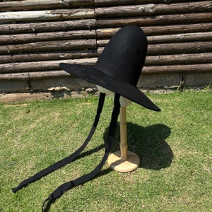 May include: A black hat with a wide brim and a tall, rounded crown. The hat features long, black ribbons. It is displayed on a wooden hat stand against a backdrop of grass and a wooden fence.