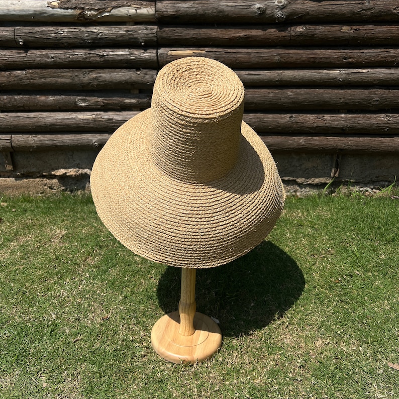 Puede incluir: Un sombrero de paja de ala ancha con una corona alta, exhibido en un soporte de madera sobre un fondo de tablones de madera y hierba verde.