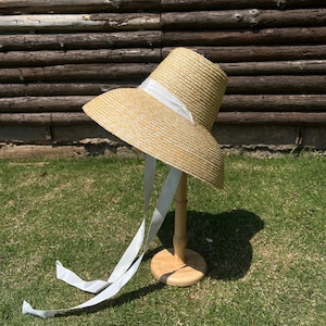 May include: A wide-brimmed straw hat with a white ribbon and long white ties. The hat is displayed on a wooden stand against a backdrop of wooden planks and green grass.
